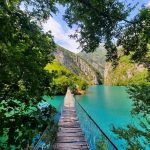 Wooden bridge crossing over the crystal-clear waters of Shala River surrounded by lush greenery.