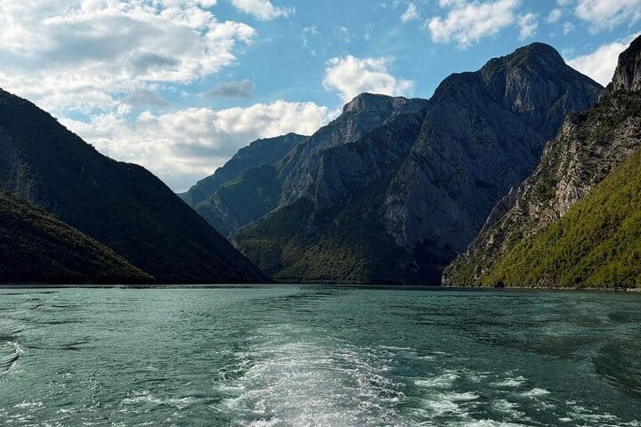 从移动的船上回望科马尼湖的宽阔景色，船尾拖着白色的水迹，两侧是高耸的岩石山峰，急剧地升起，天空布满戏剧性的云层，阿尔巴尼亚北部