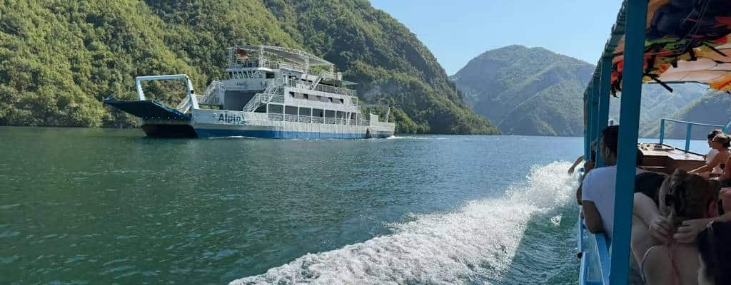 View from a small tour boat on Komani Lake as the large Alpin public ferry passes in the opposite direction, with green forested mountains behind, northern Albania