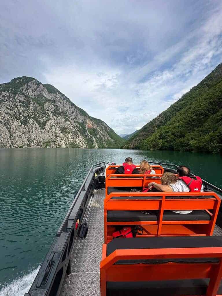 从船头看，北阿尔巴尼亚船开放式甲板的游猎船在平静的科马尼湖上，乘客穿着救生衣，戏剧性的石灰岩峡谷向前延伸至阿尔巴尼亚阿尔卑斯山。