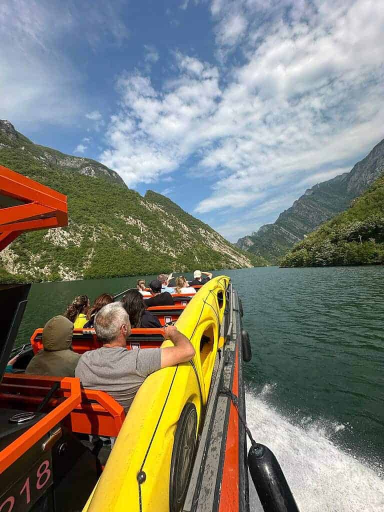 乘客们坐在阿尔巴尼亚北部奥兰治号开放式甲板的游船上，这艘船正驶过科曼湖，船的一侧绑着一条黄色的皮划艇，前方是深邃的山谷，地点是阿尔巴尼亚。