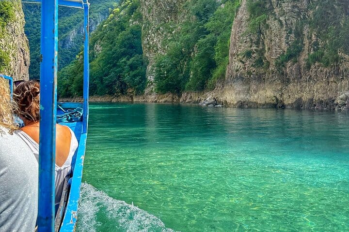 Turyści w małej niebieskiej drewnianej łodzi patrzący na krystalicznie czystą zieloną wodę i skaliste klify wąwozu rzeki Shala, okolice Jeziora Komani, północna Albania