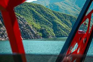 Vue à travers le cadre métallique rouge d'un navire de safari de bateau du nord de l'Albanie donnant sur l'eau turquoise du lac de Koman en direction des pentes de canyons calcaires et des crêtes montagneuses verdoyantes superposées des Alpes albanaises, Albanie