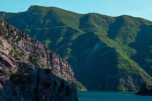 Close view of a limestone and rocky cliff on Komani Lake with densely forested mountain ridges stacked behind it and a tiny boat visible on the turquoise water below, northern Albania