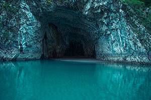A large natural limestone cave arch opening over perfectly still turquoise water on Komani Lake gorge, with the dark cave interior and a sandy floor visible in the background and the vivid blue-green water reflecting the rock walls, northern Albania