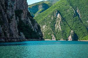 Wide view across Komani Lake showing deep turquoise water with a towering vertical limestone cliff on the left and steep densely forested mountain slopes on the right, seen during the boat crossing on the Durres to Shala River tour, Albania
