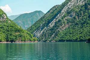 Wide view across Komani Lake looking toward the point where two forested canyon slopes converge in a V-shape, with limestone cliffs visible on the right side, calm green-blue water across the full width of the frame, and distant Albanian Alps peaks under a blue sky