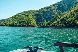 View from the bow of the North Albania Boat safari vessel showing a metal mooring cleat and bench seating in the foreground, with vivid turquoise Komani Lake water and green forested limestone canyon slopes ahead under a clear blue sky, Albania