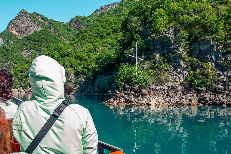 Tourist in a light hooded jacket seated on the open deck of a North Albania Boat modern vessel looking toward a white cross on a rocky cliff face above the turquoise water of Komani Lake, Albania