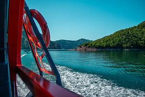 Rear view from the North Albania Boat safari vessel showing the red frame and orange life ring in the foreground, a wide white wake trailing across turquoise water, and green forested Albanian Alps canyon slopes receding into the distance under a clear blue sky