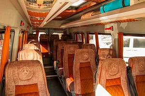 Interior of a comfortable Mercedes-Benz Sprinter minibus with embroidered brown and orange suede seats, overhead luggage racks with a rolled mat stored above, used for transfers on the North Albania Boat Shkoder to Shala River tour
