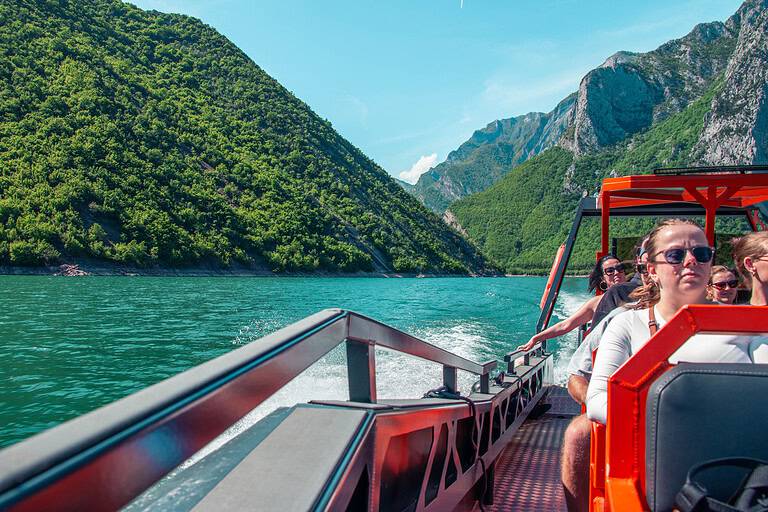 Young woman in sunglasses seated on the open orange deck of the North Albania Boat safari vessel, holding the rail as the boat speeds across the turquoise water of Komani Lake with a dramatic canyon of green mountains and limestone peaks behind her, northern Albania