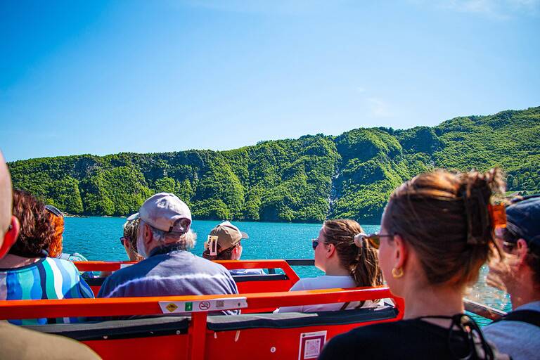 Group of mixed-age tourists seated on the open orange deck of a North Albania Boat safari vessel crossing Komani Lake under blue sky, looking out across turquoise water toward green forested canyon hills, Albania