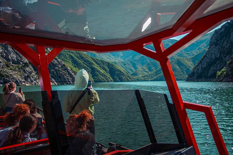 Group of tourists inside the section of a North Albania Boat private safari vessel photographing the turquoise Komani Lake canyon through the open sides, with dramatic layered green mountain ridges and limestone cliffs visible ahead, Albania