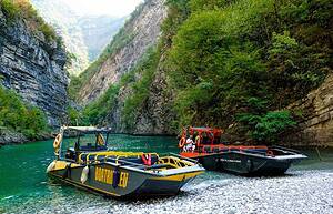 Two North Albania Boat open-deck safari vessels, one yellow and black, one orange and black, docked side by side on the gravel beach at Shala River, surrounded by towering limestone canyon walls and turquoise green water, northern Albania