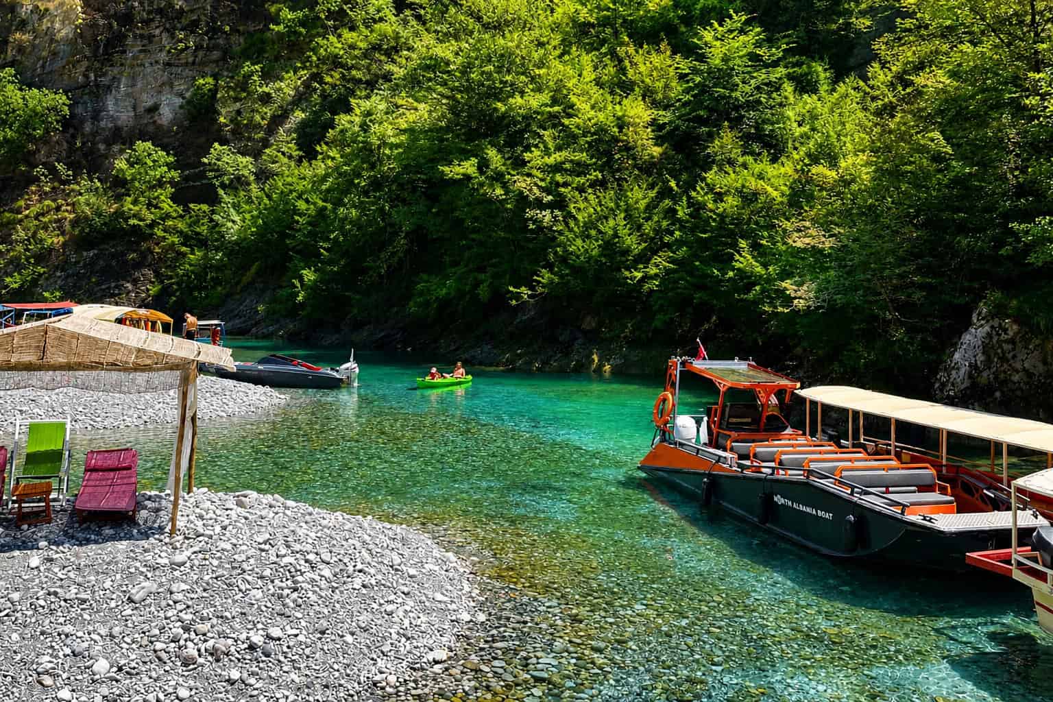 North Albania Boat orange and black safari vessel docked at the gravel Shala River beach alongside kayakers on crystal clear turquoise water, with straw sun shelters, sun loungers and green canyon walls visible, northern Albania