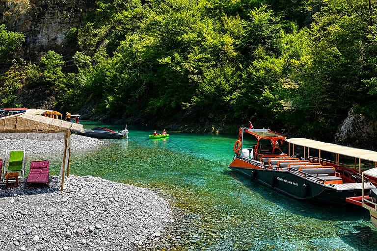 North Albania Boat orange and black safari vessel docked at the gravel Shala River beach alongside kayakers on crystal clear turquoise water, with straw sun shelters, sun loungers and green canyon walls visible, northern Albania