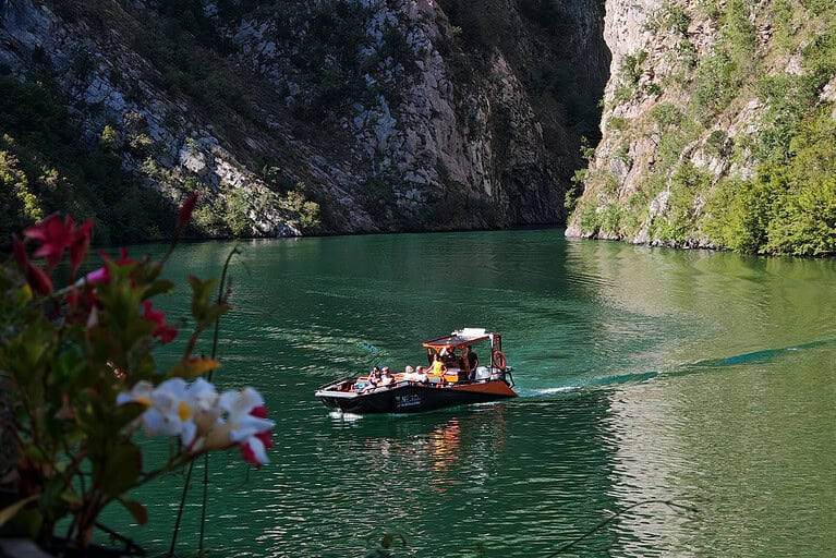 North Albania Boat orange and black safari vessel with passengers crossing the deep green water of the Shala River gorge, framed in the foreground by red and white wildflowers, with limestone canyon walls rising on both sides, Albania