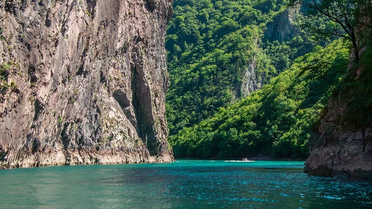 Boat on turquoise water between towering limestone cliffs and forested green slopes in the Komani–Shala canyon, northern Albania boat tour