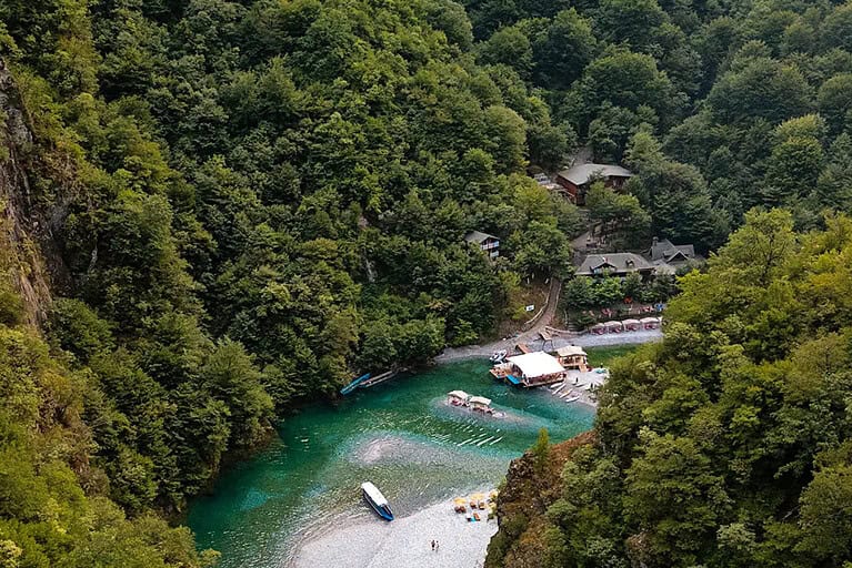 Aerial view looking down into the Shala River canyon showing the turquoise river, gravel beach, floating restaurant platform, rowboats, sun loungers, and wooden buildings tucked into the forest, with limestone canyon walls on the left, northern Albania