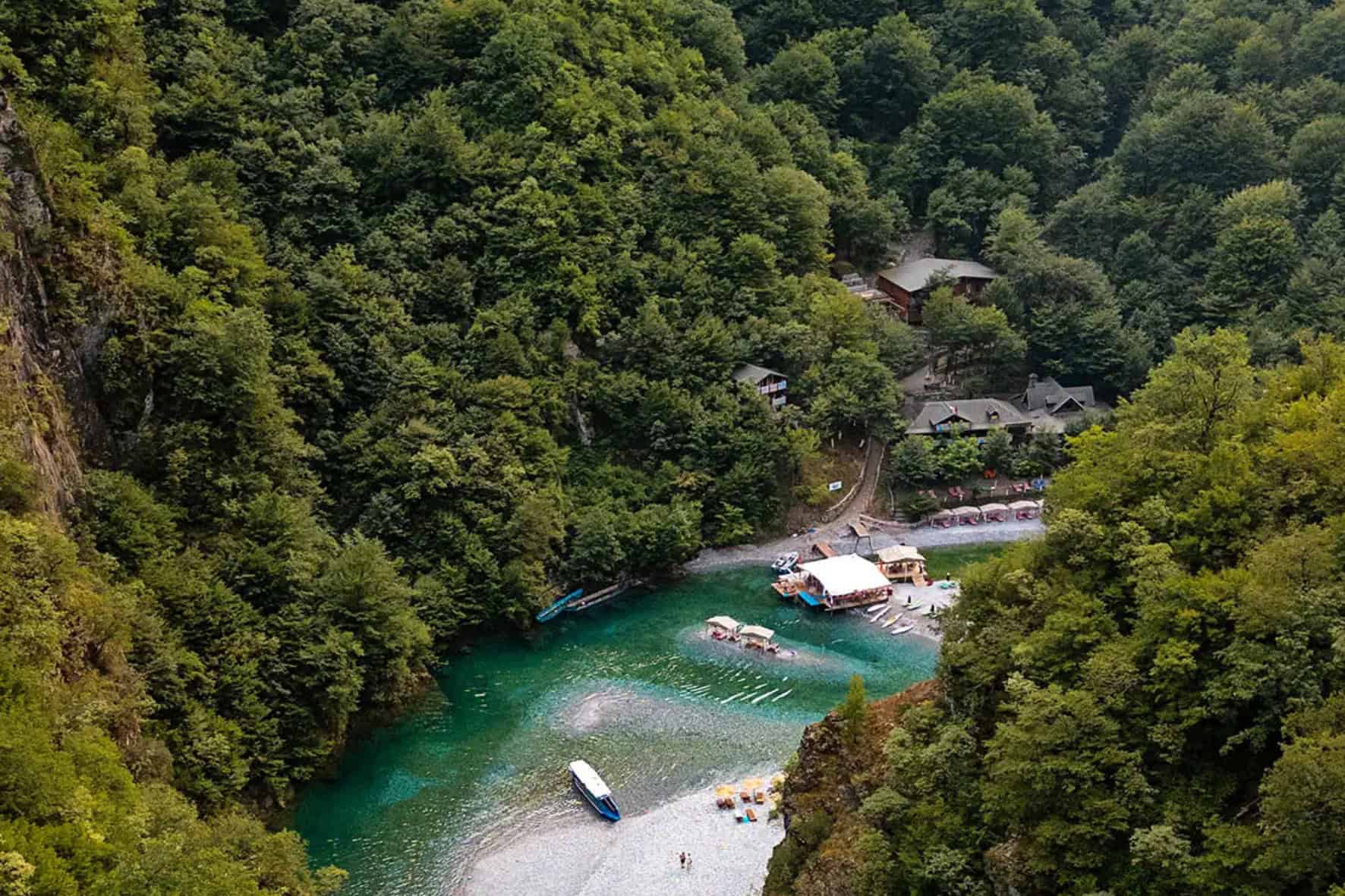 Aerial view looking down into the Shala River canyon showing the turquoise river, gravel beach, floating restaurant platform, rowboats, sun loungers, and wooden buildings tucked into the forest, with limestone canyon walls on the left, northern Albania