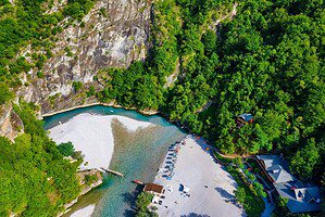 Aerial drone photograph looking down at the full Shala River beach showing the white gravel shoreline, turquoise river, sun loungers and umbrellas, restaurant buildings, and the inner canyon mouth, surrounded by dense green forest and limestone cliffs, northern Albania