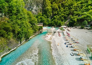 Aerial view of the Shala River beach showing rows of colourful sun loungers and straw umbrellas along the wide gravel shore, vivid turquoise river water with a wooden footbridge, and steep green forested canyon walls on both sides, northern Albania