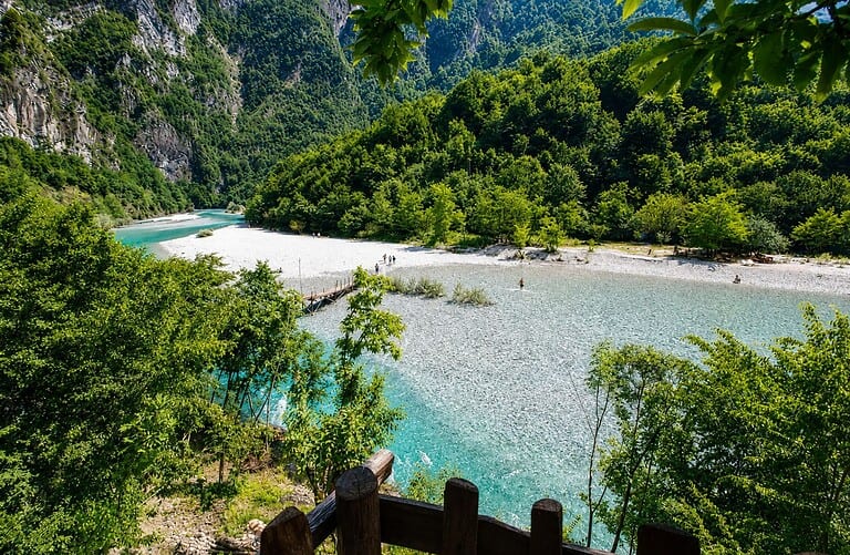 View from an elevated wooden railed terrace looking down over the wide Shala River beach, turquoise water, wooden footbridge, and narrow inner canyon entrance surrounded by dense green forest and limestone cliffs, northern Albania