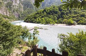 View from an elevated wooden railed terrace looking down over the wide Shala River beach, turquoise water, wooden footbridge, and narrow inner canyon entrance surrounded by dense green forest and limestone cliffs, northern Albania