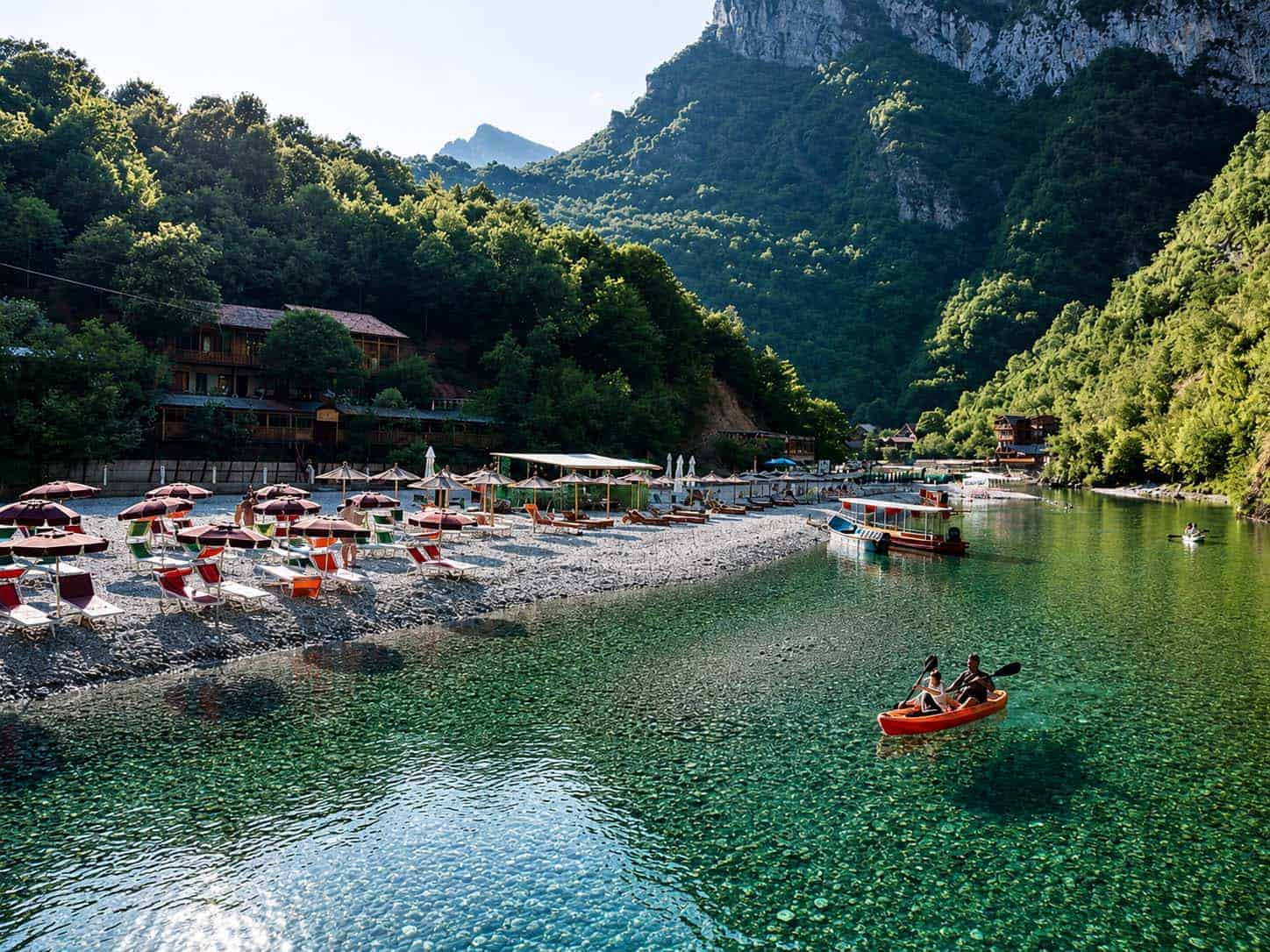 Two people in an orange kayak on turquoise Shala River water, with a pebble beach full of sun loungers and umbrellas and forested Albanian Alps mountains behind