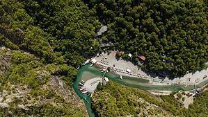 High aerial drone view looking down at the full Shala River beach in peak summer season, showing rows of sun loungers and umbrellas, multiple boats docked along the turquoise river, restaurant buildings, and dense green canyon forest on all sides, northern Albania