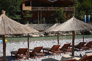 Rows of wooden sun loungers and natural straw umbrellas on the Shala River gravel beach in front of a two-storey wooden restaurant building with a balcony, with the turquoise river and stone wall visible behind, northern Albania