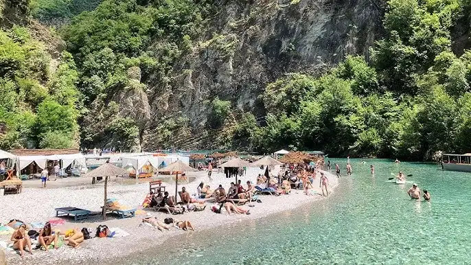 Busy Shala River pebble beach with sunbathers under thatched parasols and swimmers in turquoise water beneath limestone canyon cliff, northern Albania