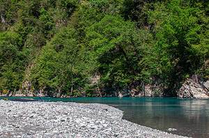 Wide white and grey gravel beach at Shala River with a wooden rowboat and folding sun lounger at the water's edge, calm turquoise river water and lush green forested canyon walls in the background, northern Albania