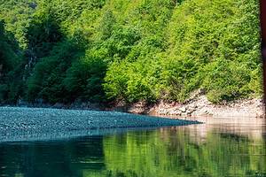 View from the North Albania Boat safari vessel looking toward a smooth grey gravel bank on Komani Lake, with vivid green spring foliage and its perfect mirror reflection in the still turquoise-green water, northern Albania