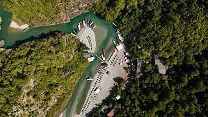 Direct top-down aerial drone photograph of the Shala River showing the turquoise river bend, multiple boats fanned out on the gravel beach, rows of umbrellas, restaurant structures in the forest, and the inner canyon visible to the left, northern Albania