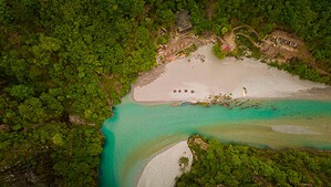 Vue aérienne directe du dessus de la rivière Shala montrant une large plage de sable et de gravier blanc qui ondule à côté d'une eau d'un turquoise vif, avec des kayaks colorés sur le rivage, des structures en bois et une forêt verte dense de tous côtés, nord de l'Albanie