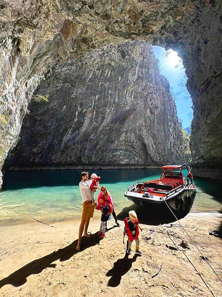 Family with children standing inside a large natural limestone cave on the Shala River, with a red and black tour boat moored on the sandy shore and turquoise water visible through the cave opening