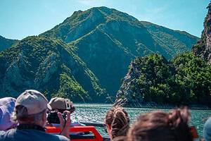 Turisti seduti sul ponte aperto di una moderna imbarcazione dell'Albania settentrionale che fotografano il drammatico canyon roccioso e l'acqua turchese del lago di Koman, Albania