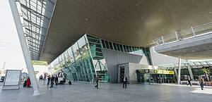 Extérieur du terminal des arrivées de l'aéroport international de Tirana montrant la façade moderne en verre et en acier, la signalisation jaune des arrivées en albanais et en anglais, et des passagers avec des bagages sur l'aire de stationnement, Albanie