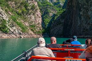 Tourists seated on the open orange deck of a North Albania Boat safari vessel looking forward into the dramatic narrowing gorge of Komani Lake, with towering limestone cliff faces and turquoise water visible ahead, northern Albania