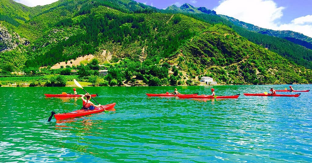 Group of tourists paddling red kayaks across turquoise Komani Lake with green mountain slopes and village on far shore, northern Albania