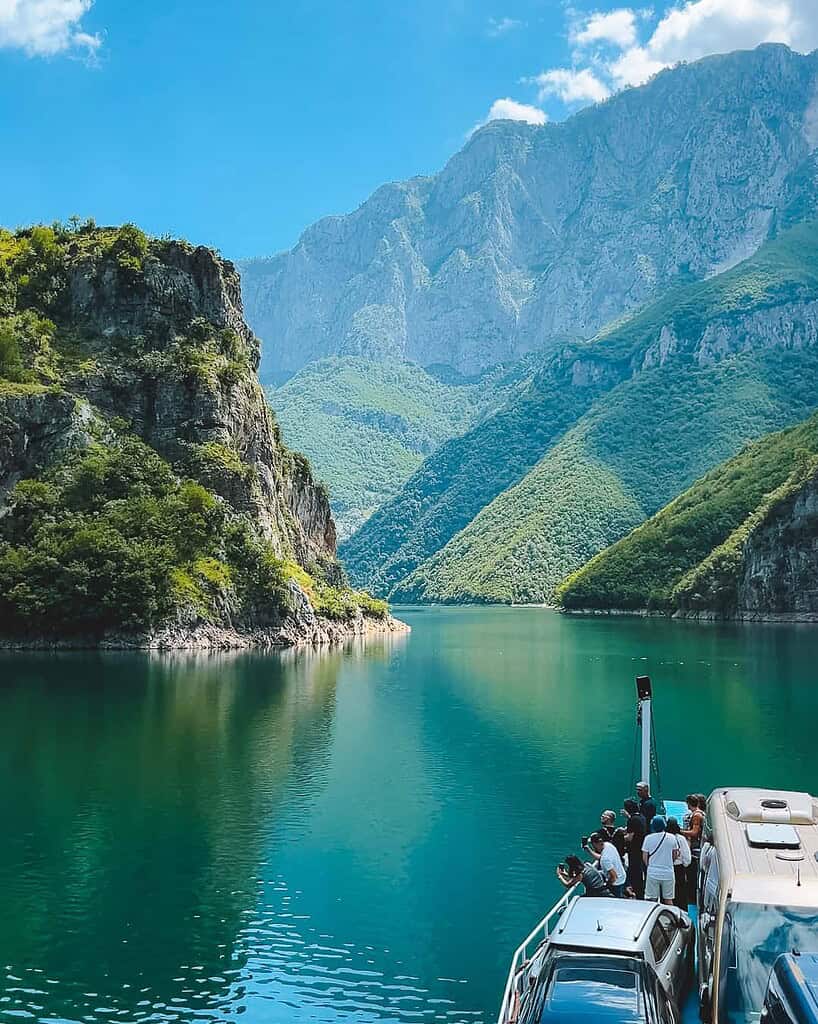 Tourists on boat deck photographing Komani Lake canyon with mirrored green water and Albanian Alps peaks, northern Albania