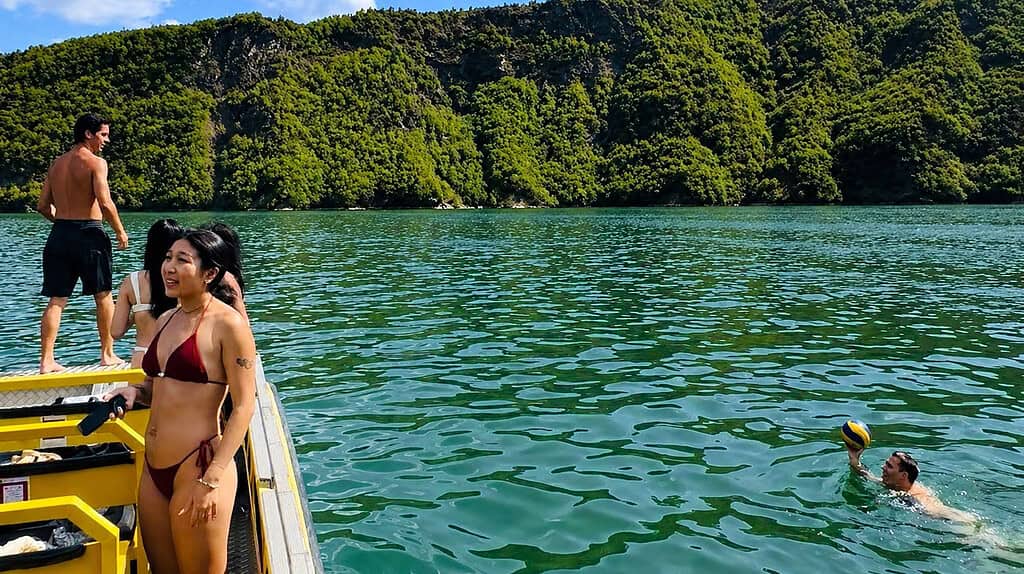 People swimming from a boat on Komani Lake in Albania with green mountain cliffs and clear turquoise water.