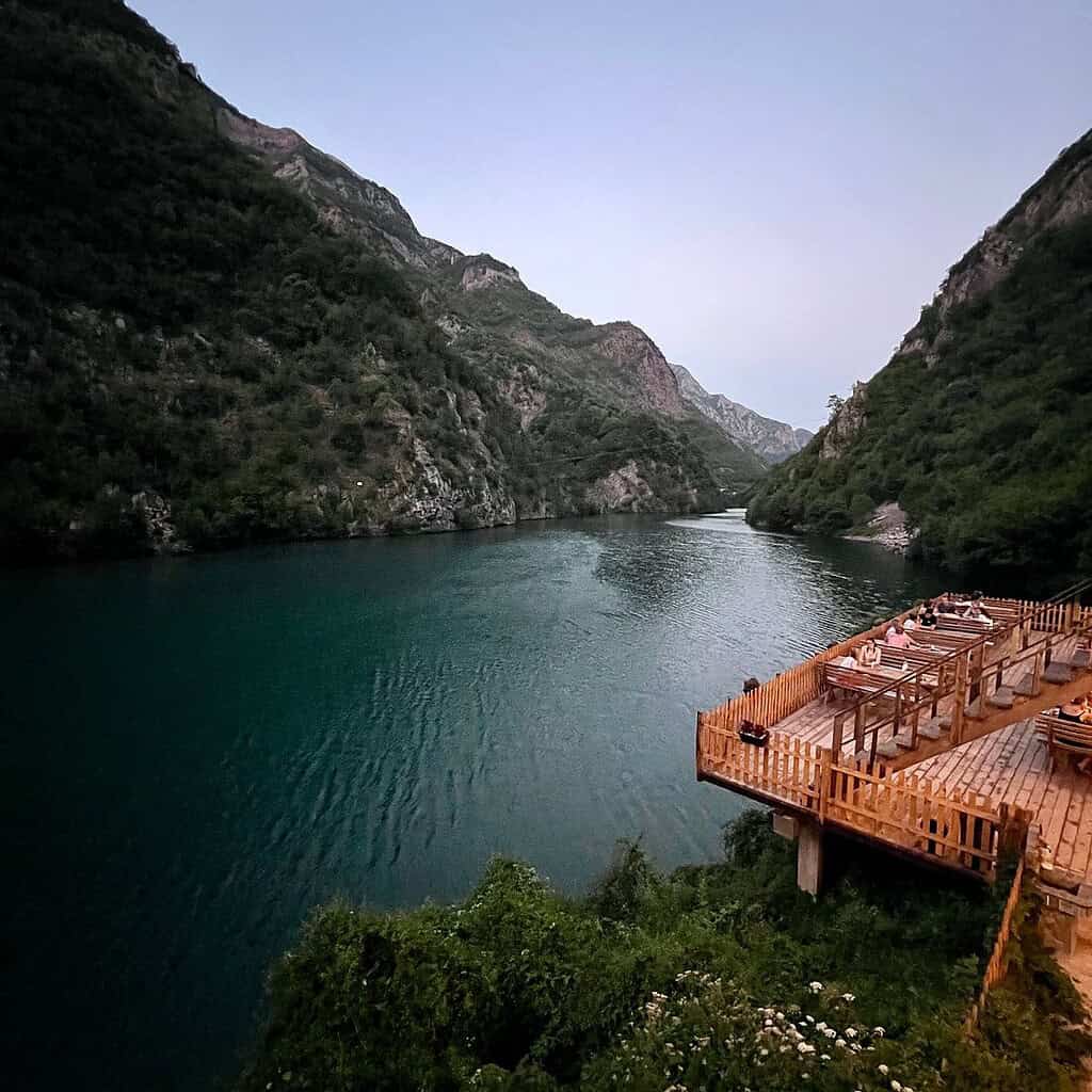 Guests on wooden overwater deck at dusk overlooking deep blue-green Komani Lake canyon and mountain silhouettes, northern Albania