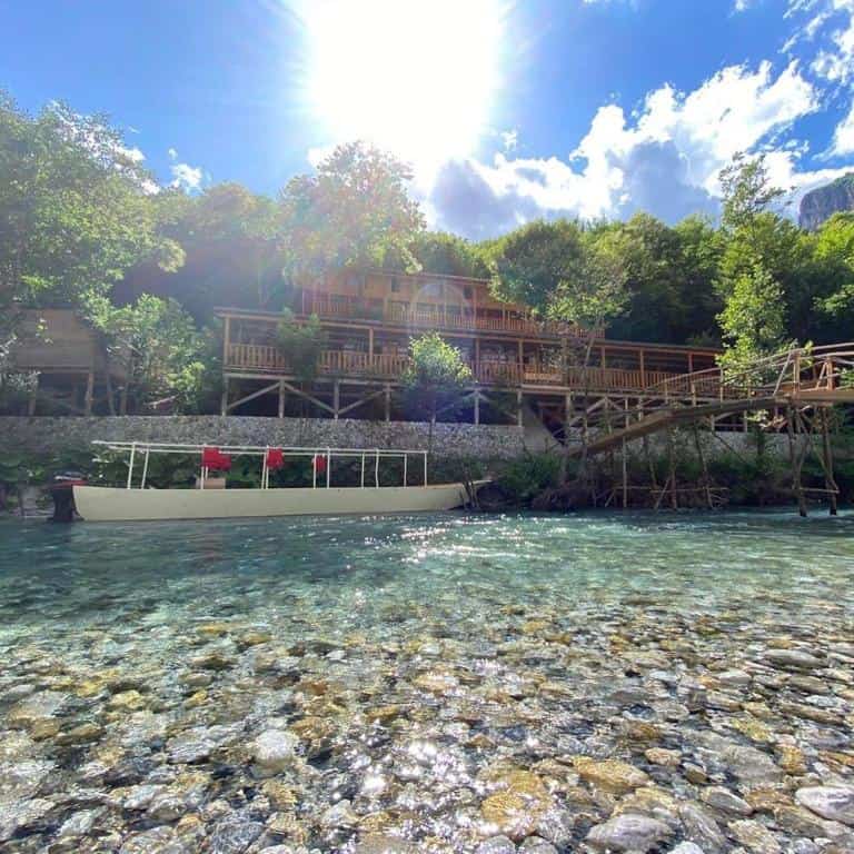 Timber waterfront lodge above crystal clear Komani Lake shallows with white boat moored at dock, northern Albania