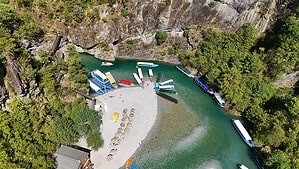 Aerial view of Shala River Beach in Albania with turquoise water, tourist boats, kayaks, pebble beach, and the limestone canyon on Komani Lake.