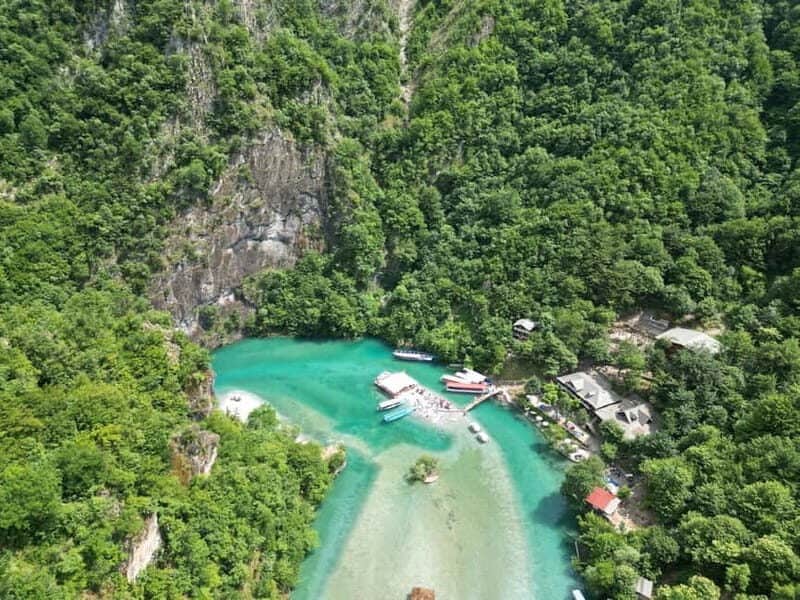High aerial view of Shala River canyon with emerald water, beach, moored boats and forested limestone walls, northern Albania