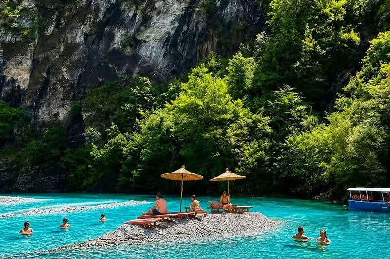 Tourists relaxing on pebble island sun loungers in the middle of turquoise Shala River beneath canyon cliffs, northern Albania