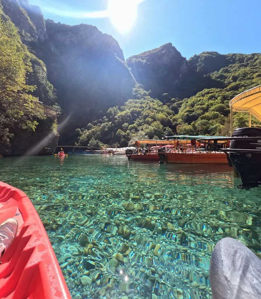 View from a kayak on the clear water of Shala River with boats in the background, Albania